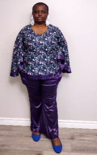 Plus size woman wearing a floral blouse and purple pants against a white wall.