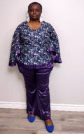 Plus size woman wearing a floral blouse and purple pants against a white wall.