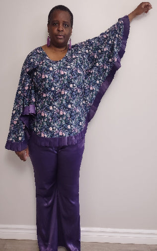 Plus size woman wearing a floral blouse with purple trim and matching pants against a white wall.
