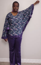 Plus size woman wearing a floral blouse with purple trim and matching pants against a white wall.
